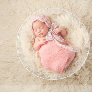 May include: A newborn baby girl sleeping in a white wire basket lined with a fluffy white blanket. The baby is wearing a pink knitted hat with a white floral ribbon and a pink swaddle.
