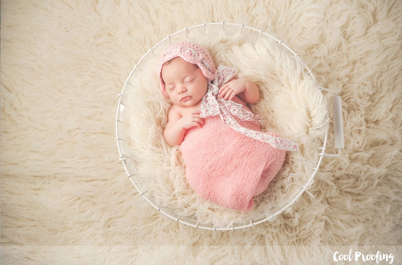May include: A newborn baby girl sleeping in a white wire basket lined with a fluffy white blanket. The baby is wearing a pink knitted hat with a white floral ribbon and a pink swaddle.