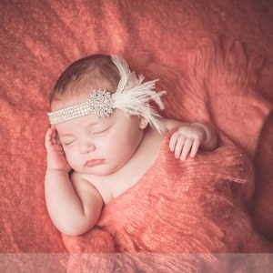 May include: A sleeping baby girl wearing a white feather headband with rhinestones on a coral colored blanket.