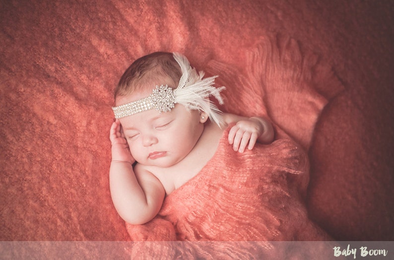 May include: A sleeping baby girl wearing a white feather headband with rhinestones on a coral colored blanket.
