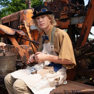 May include: A young person wearing a brown shirt, a black bowler hat with goggles, and a white and blue patchwork apron, sits on a rusty metal structure. The person is holding a small metal object in their hands.