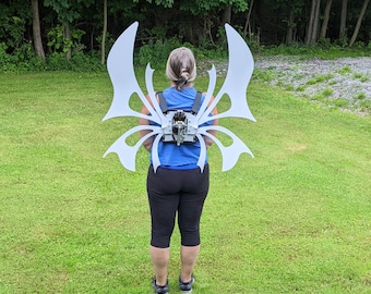 May include: A person wearing a blue shirt and black pants with a white, intricate, butterfly-shaped wing costume attached to their back.