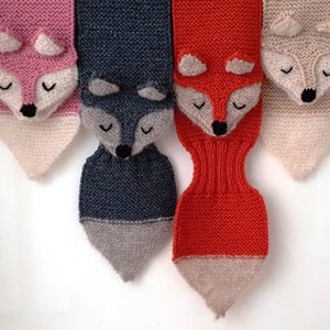 Four fox scarves hanging side by side. Each handknitted in a diffrent color of merino wool. Pink, dark gray, burnt orange and beige.