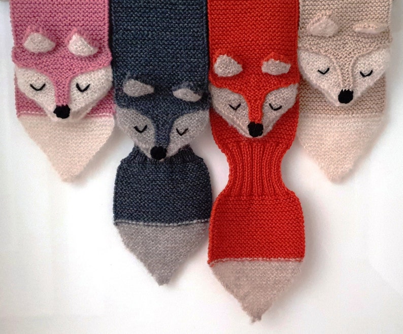 Four fox scarves hanging side by side. Each handknitted in a diffrent color of merino wool. Pink, dark gray, burnt orange and beige.