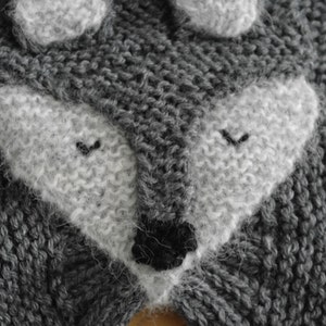 May include: Close-up of a gray knitted fabric with a white knitted wolf face. The wolf has a black nose and closed eyes.