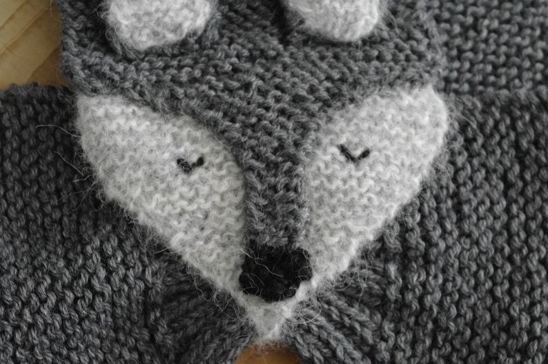 May include: Close-up of a gray knitted fabric with a white knitted wolf face. The wolf has a black nose and closed eyes.