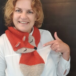 May include: A woman wearing a white shirt is pointing to a knitted fox scarf. The scarf is orange and beige with a fox face on the front.