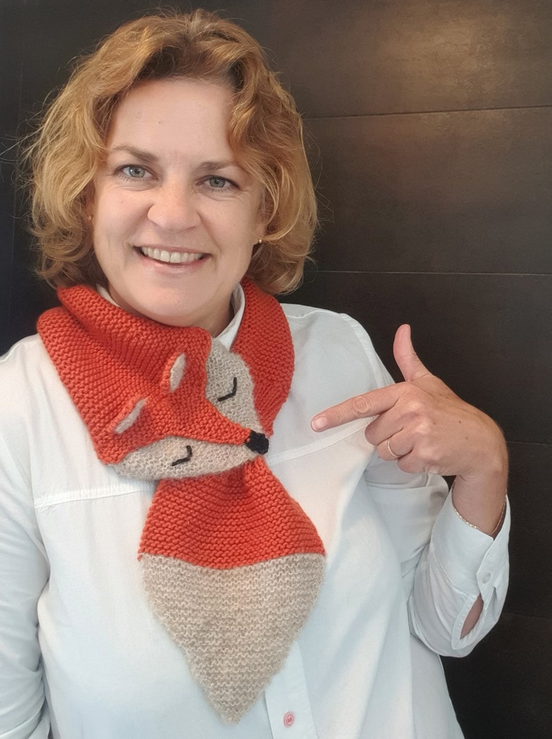 May include: A woman wearing a white shirt is pointing to a knitted fox scarf. The scarf is orange and beige with a fox face on the front.