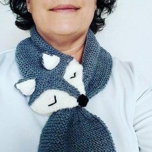 May include: A gray knitted scarf with a white fox face appliqu&eacute;. The fox has black eyes and a black nose.