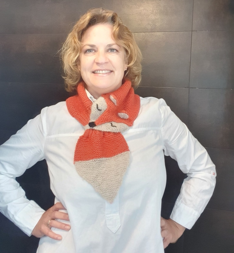 May include: A woman wearing a white button-down shirt with a knitted fox scarf. The scarf is orange and beige with a fox face design.