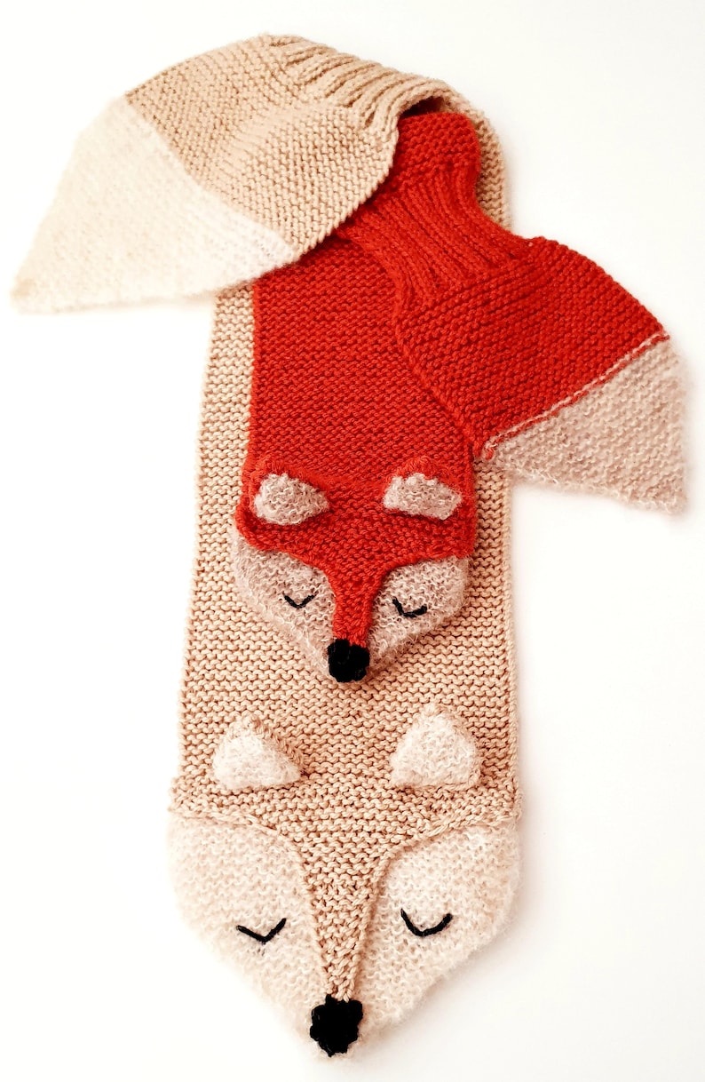 May include: A knitted scarf with two fox faces, one orange and one beige. The scarf is made with a soft, fluffy yarn.