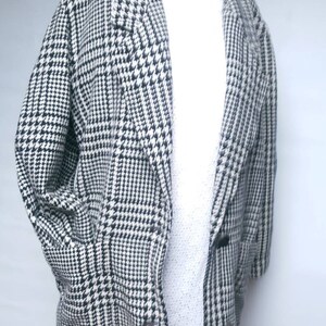 May include: A black and white houndstooth patterned coat with a long, loose fit. The coat has a collar and a button closure.