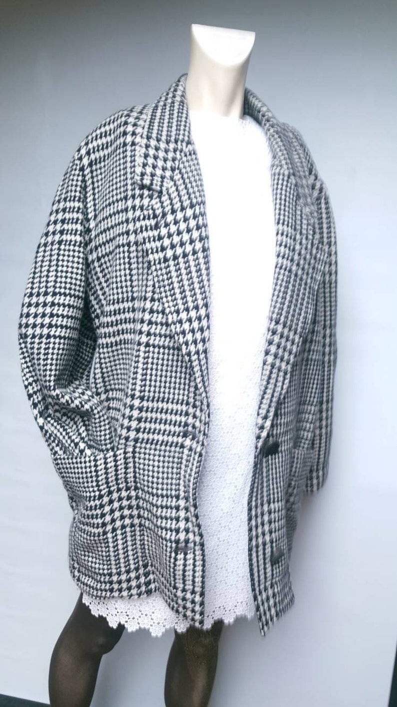 May include: A black and white houndstooth patterned coat with a long, loose fit. The coat has a collar and a button closure.