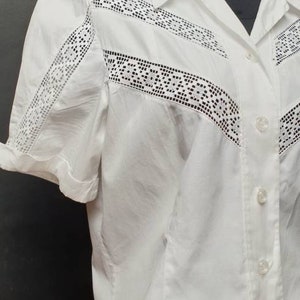 May include: A white short-sleeved button-up shirt with a lace trim. The lace trim is black and has a geometric pattern.