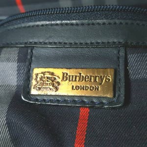 Puede incluir: Un bolso de cuero negro con una etiqueta dorada de Burberrys London. El bolso tiene un patrón de cuadros con rayas negras, blancas y rojas.
