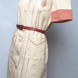 Puede incluir: Un vestido de color crema con un cinturón rojo y un velero bordado en el pecho. El vestido tiene cuello y mangas cortas con ribetes rosas.