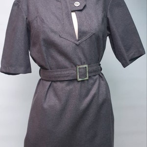 Puede incluir: Un vestido gris de manga corta con cuello alto y escote en V. El vestido tiene un cinturón con una hebilla plateada.