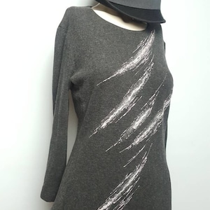 Puede incluir: Un vestido gris oscuro de manga larga con un diseño abstracto blanco. El vestido tiene un escote redondo y una silueta ajustada. Un sombrero fedora negro está encima del vestido.