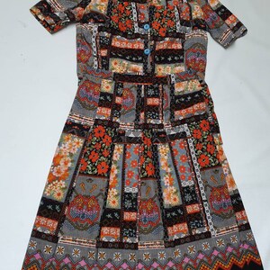 May include: A vintage two-piece dress with a short-sleeved top and a long skirt. The dress is made of a black fabric with a colourful patchwork pattern featuring red, orange, yellow, green, blue, and white flowers. The top has a button-up closure and a collar.