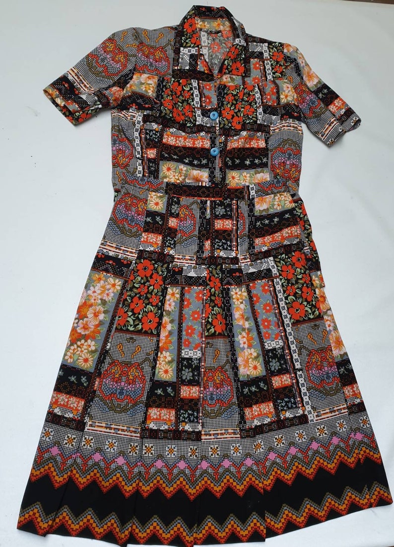 May include: A vintage two-piece dress with a short-sleeved top and a long skirt. The dress is made of a black fabric with a colourful patchwork pattern featuring red, orange, yellow, green, blue, and white flowers. The top has a button-up closure and a collar.