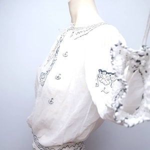 May include: White cotton blouse with black embroidery. The blouse has a gathered neckline and a ruffled sleeve. The embroidery features a floral design and a geometric pattern.