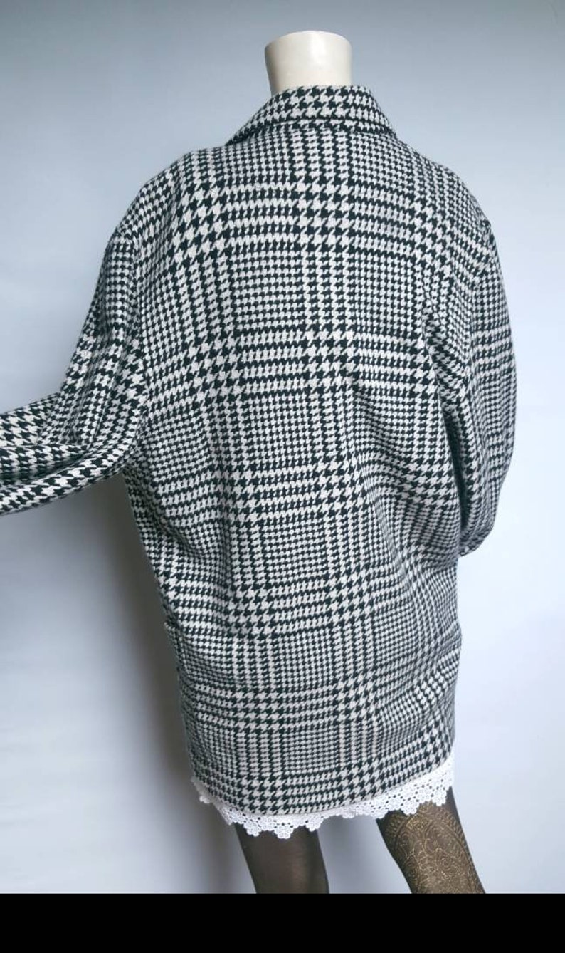 May include: Black and white houndstooth patterned coat with a long sleeve and a collar.