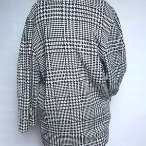 May include: A black and white houndstooth patterned jacket with long sleeves and a collar. The jacket is on a mannequin and is worn over a white lace skirt.