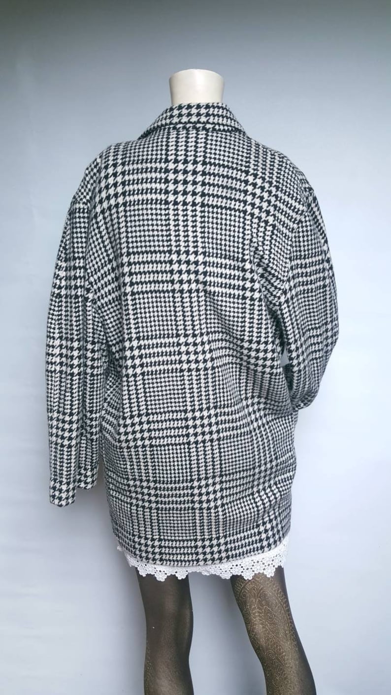 May include: A black and white houndstooth patterned jacket with long sleeves and a collar. The jacket is on a mannequin and is worn over a white lace skirt.