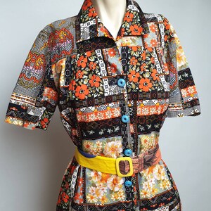 May include: A short-sleeved button-up shirt with a patchwork design featuring orange, black, and white floral patterns. The shirt has a collar and a yellow belt with a brown buckle.