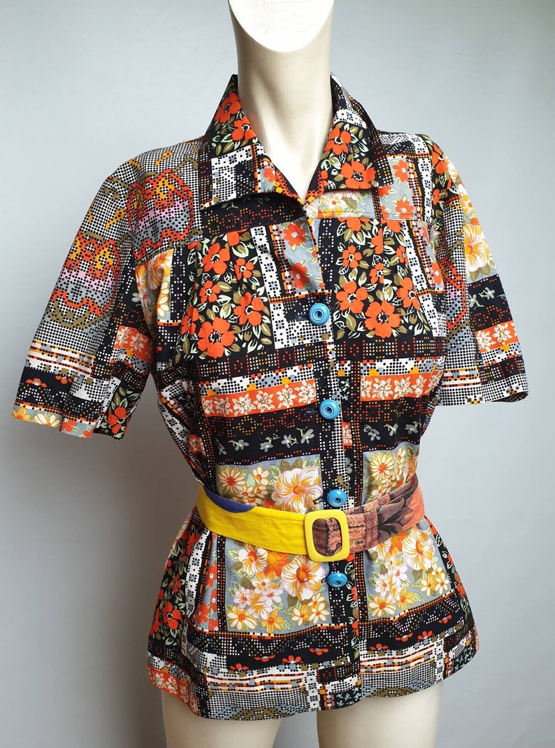 May include: A short-sleeved button-up shirt with a patchwork design featuring orange, black, and white floral patterns. The shirt has a collar and a yellow belt with a brown buckle.