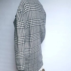May include: A black and white houndstooth patterned jacket with a long sleeve and a collar. The jacket is on a mannequin and the bottom of the jacket is cropped above the knees.