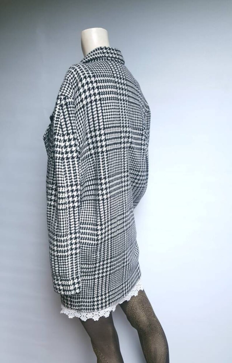 May include: A black and white houndstooth patterned jacket with a long sleeve and a collar. The jacket is on a mannequin and the bottom of the jacket is cropped above the knees.