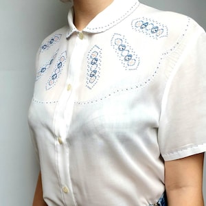 Puede incluir: Camisa blanca de manga corta con cuello de Peter Pan y detalles bordados en azul y rosa.