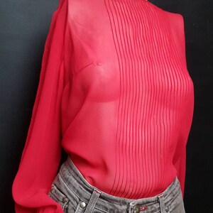 Puede incluir: Una blusa roja transparente con mangas largas y un frente plisado. La blusa se lleva sobre un pantalón vaquero gris.