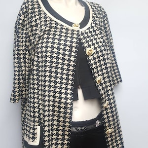 May include: A black and white houndstooth patterned cardigan with gold buttons and a black trim. The cardigan has a scoop neck and short sleeves.