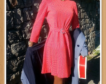 Puede incluir: Un vestido rojo con lunares blancos y un cinturón negro. El vestido tiene una falda acampanada y mangas largas. El maniquí lleva una chaqueta azul sobre el vestido.