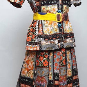 May include: A vintage two-piece outfit featuring a short-sleeved blouse and a skirt. The fabric is a black and white patchwork with orange and yellow floral patterns. The blouse has a collar and buttons. The skirt has a yellow belt with a blue buckle.