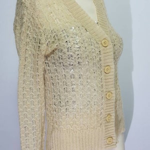 May include: A beige knitted cardigan with a V-neck and button closure. The cardigan is made of a lightweight, open-weave fabric.