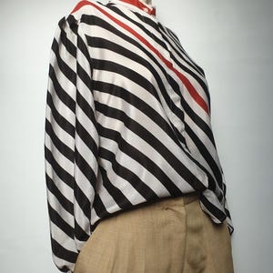 May include: A long-sleeved blouse with black and white stripes and a red accent. The blouse has a high collar with a red trim and is paired with tan-colored pants. The image is taken from a side angle.