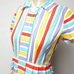 Puede incluir: Un vestido de rayas de colores con cuello blanco y mangas cortas. El vestido tiene cierre de botones y una falda plisada.