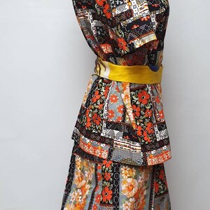 May include: A black and orange patchwork print dress with a yellow belt. The dress has a short-sleeved top and a long skirt with a tiered design.