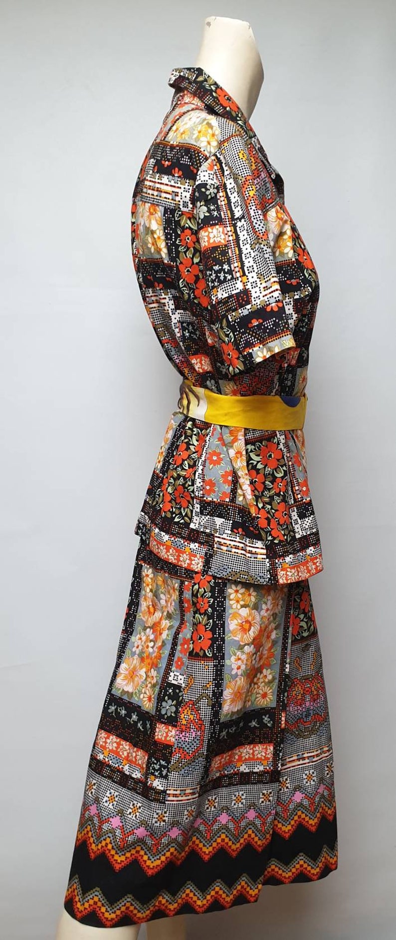 May include: A black and orange patchwork print dress with a yellow belt. The dress has a short-sleeved top and a long skirt with a tiered design.