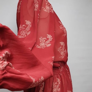 Pode incluir: Um vestido maxi vermelho transparente com estampa floral branca. O vestido tem cintura franzida e mangas compridas.