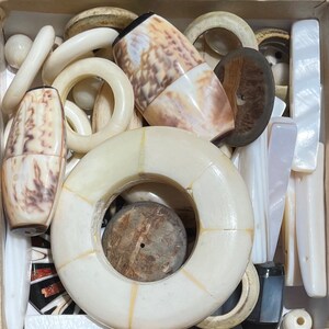 Puede incluir: Una colección de piezas de joyería de concha de color blanco, marrón y crema, incluyendo anillos, colgantes y cuentas. Las piezas están dispuestas en una caja, y algunas están apiladas unas sobre otras.