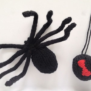 May include: A black knitted spider with eight legs and a red hourglass on its abdomen. The spider is hanging from a black string.