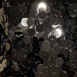 May include: A close-up image of a large quantity of black round sequins. The sequins are shiny and have a metallic finish.