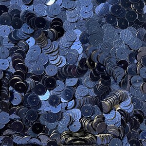 Puede incluir: Primer plano de un montón de lentejuelas azul marino. Las lentejuelas son redondas con un pequeño agujero en el centro y tienen una superficie brillante y reflectante. Las lentejuelas están dispersas y superpuestas, creando una apariencia texturizada.