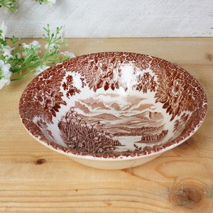 Può includere: Una ciotola in ceramica bianca con un motivo floreale marrone. La ciotola presenta una scena di paesaggio con montagne, alberi e un fiume.