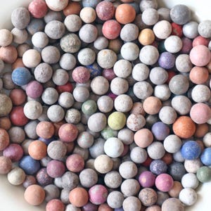 Puede incluir: Una colección de pequeñas canicas redondas y coloridas en un cuenco blanco. Las canicas varían en color, incluyendo tonos de rosa, azul, naranja, verde y gris. Algunas canicas tienen un aspecto moteado, mientras que otras son de color sólido. El cuenco está lleno de una variedad de tamaños.