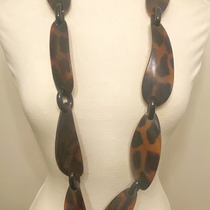 Peut inclure: Un long collier brun et noir compos&eacute; de grandes pi&egrave;ces ovales. Les pi&egrave;ces sont reli&eacute;es par des anneaux m&eacute;talliques noirs.